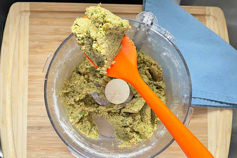 Horizontal image of a light green paste in a food processor with an orange spatula.