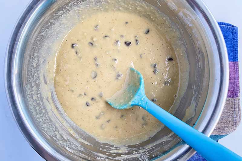 Horizontal image of a wet mixture studded with chocolate chips with a blue spatula in a metal bowl.