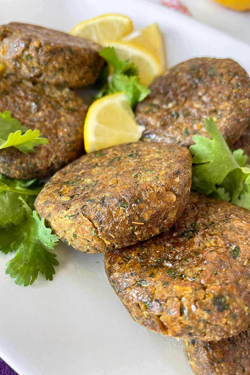 Vertical close-up image of small chickpea patties on a plate with herbs and lemon wedges.