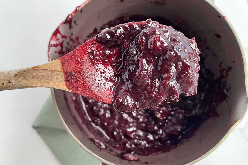 Horizontal image of a thickened blueberry spread mixture in a pot with a wooden spoon.