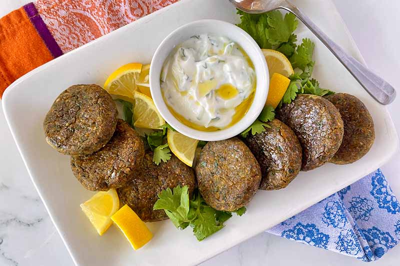 Horizontal image of a platter of falafel with lemon wedges, herbs, and a bowl of dip.
