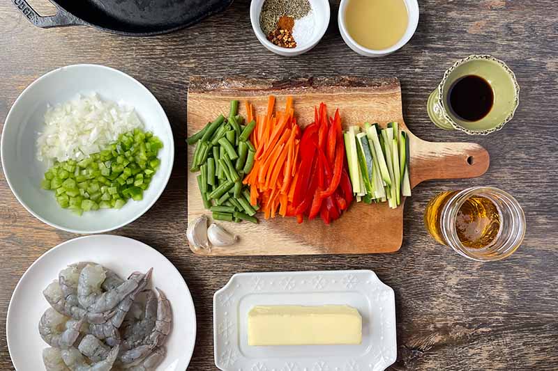 Horizontal image of prepped vegetables, shrimp, and assorted seasonings.
