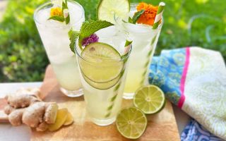 Horizontal image of glasses filled with a light beverage with straws garnished with lime, herbs, and flowers on a wooden board next to citrus and a colorful towel.