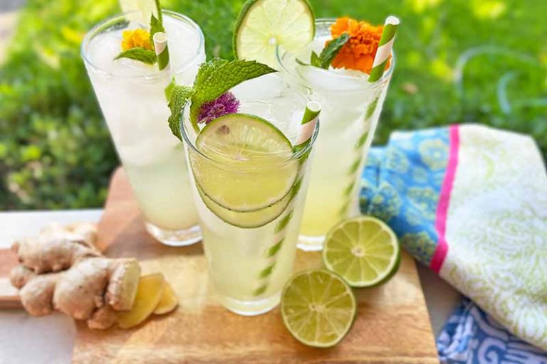 Horizontal image of glasses filled with a light beverage with straws garnished with lime, herbs, and flowers on a wooden board next to citrus and a colorful towel.
