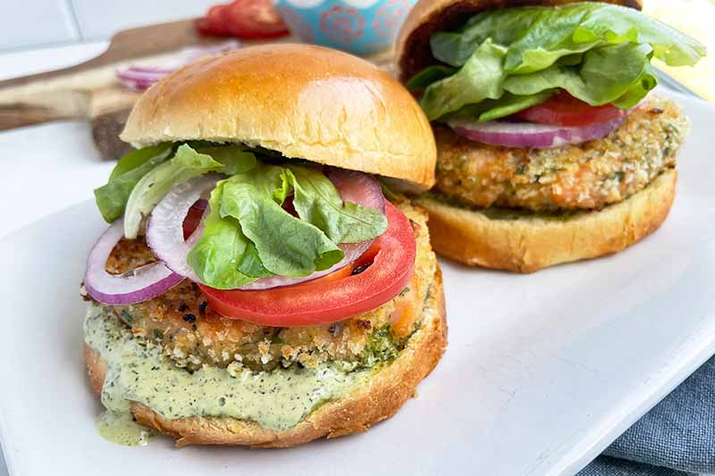 Horizontal image of two sandwiches with seafood patties with an herb sauce, tomatoes, lettuce, and red onions.