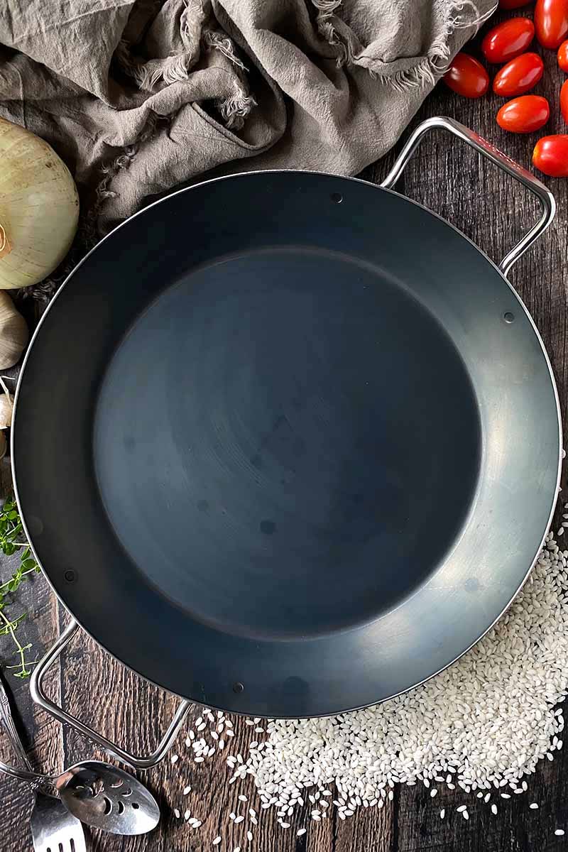 Vertical top-down image of an empty circular piece of cookware with silver handles next to rice, a tan towel, and fresh produce.