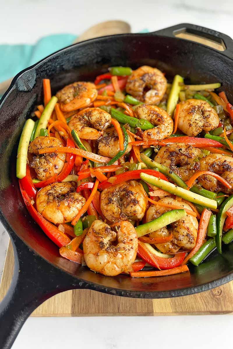 Vertical image of a cast iron pan filled with sliced bell peppers, zucchini, and seafood.