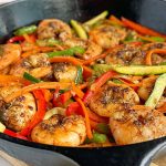 Horizontal image of a cast iron pan filled with cooked sliced zucchini, red bell peppers, and shrimp.