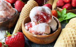 Horizontal image of a wooden bowl filled with scoopfuls of a frozen dessert next to fresh fruit and green herbs.