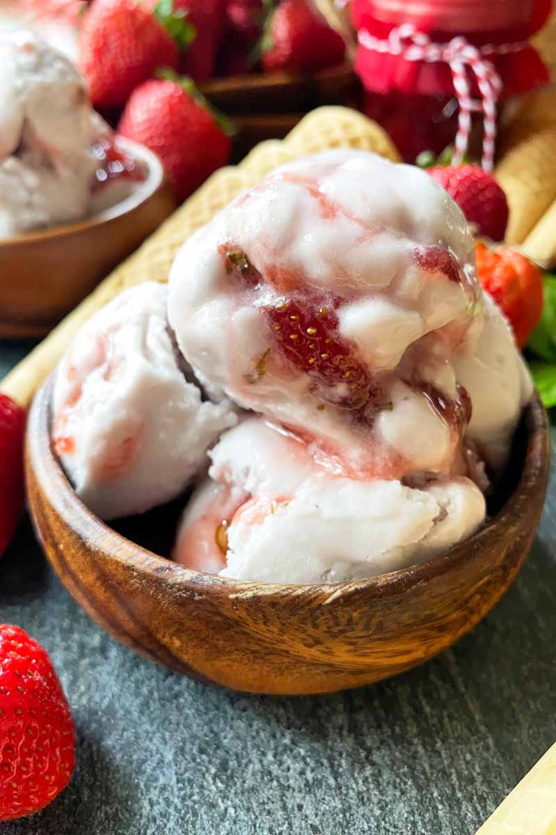 Vertical close-up image of a wooden bowl filled with scoopfuls of a frozen dessert next to fresh fruit.