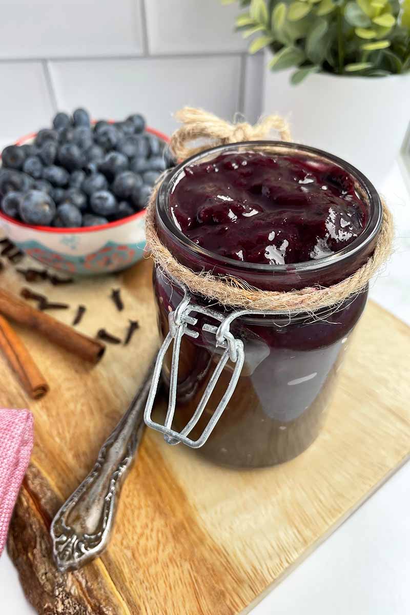 Vertical image of a jar filled with a dark purple fruit compote next to a bowl of fruit on a wooden cutting board.