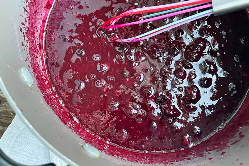 Horizontal image of whisking a dark purple fruit compote mixture in a pot.