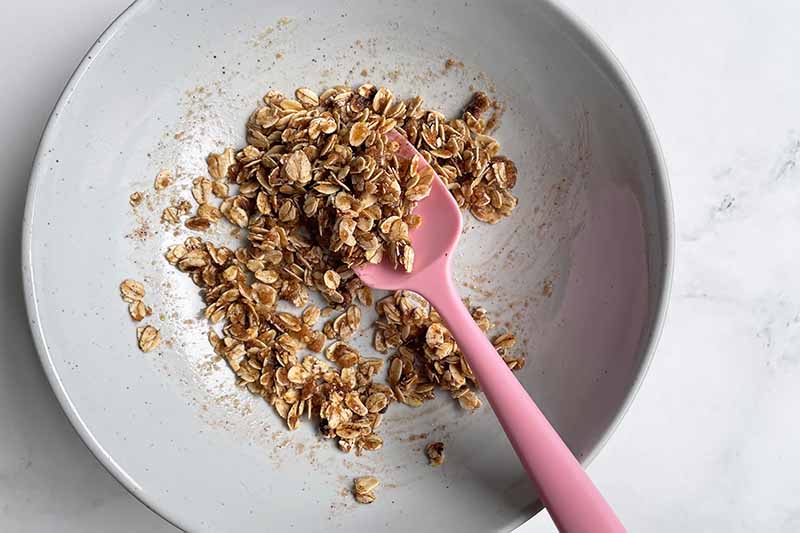 Horizontal image of making an oat streusel topping in a bowl mixed with a pink spatula.
