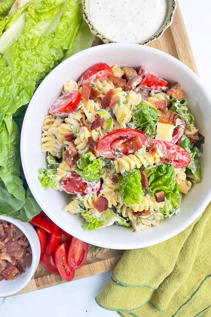 Vertical top-down image of a white bowl with a pasta salad next to lettuce, tomatoes, bacon in a bowl, ranch dressing in a bowl, and a green towel.