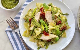 Horizontal image of a white dinner plate with farfalle mixed with chicken slices and assorted vegetables in a green sauce next to a fork and napkin.