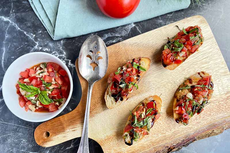 Horizontal image of bruschetta drizzled with vinegar and chopped with herbs on a wooden cutting board.