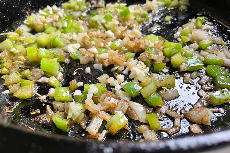Horizontal image of cooking celery, green bell peppers, onion, and garlic in a cast iron pan. 