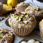 Horizontal image of individual lined baked goods topped with chopped nuts and sanding sugar on a pan next to lemons.