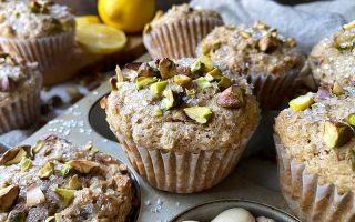 Horizontal image of individual lined baked goods topped with chopped nuts and sanding sugar on a pan next to lemons.