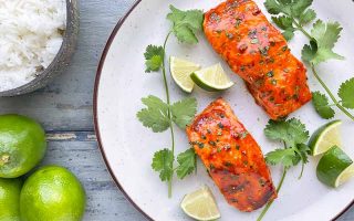 Horizontal image of two fillets of seasoned and cooked fish on a white plate with herbs and lime wedges.