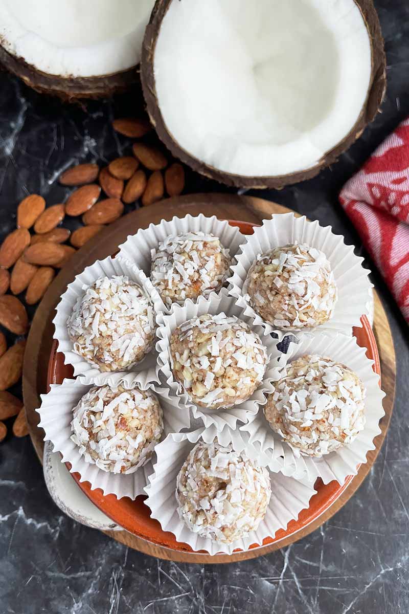 Vertical image of small snacks in white paper muffin liners on a plate next to almonds and a halved fresh coconut.