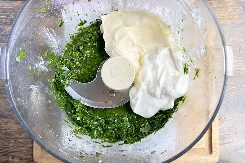 Horizontal image of a dollop of mayonnaise and sour cream on top of chopped herbs in a food processor.