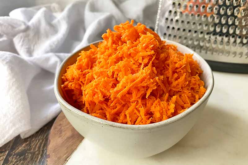 Horizontal image of finely shredded carrots in a white bowl in front of a metal kitchen appliance.
