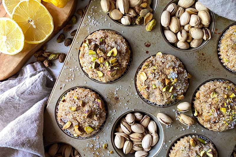 Horizontal top-down image of individual baked goods topped with chopped nuts and sugar surrounded by whole nuts, sugar, and slices of lemons.