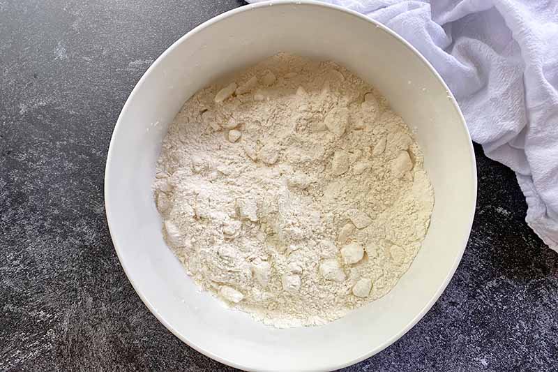Horizontal image of a flour mixture with chunks of fat in a large white bowl.