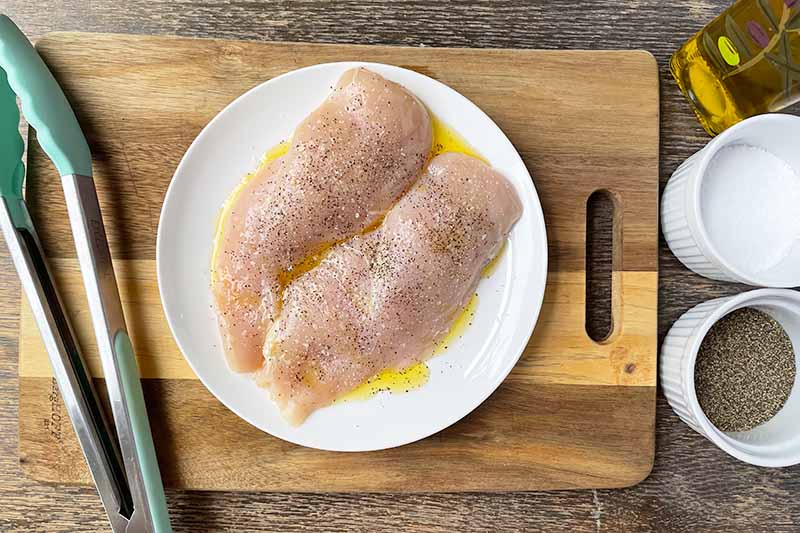 Horizontal image of raw poultry breasts lightly seasoned on a white plate next to tongs on a wooden cutting board.