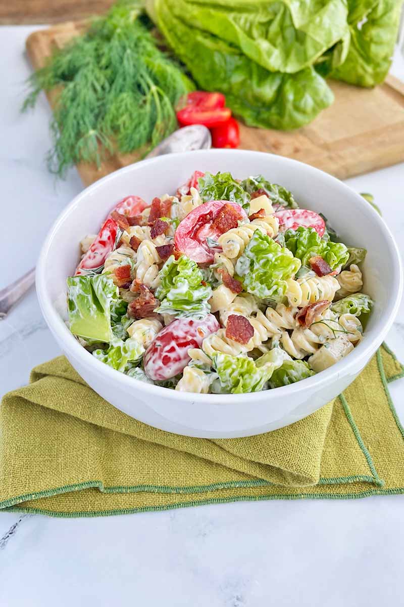 Vertical image of a white bowl filled with rotini, shredded lettuce, and fresh tomatoes on a green towel.