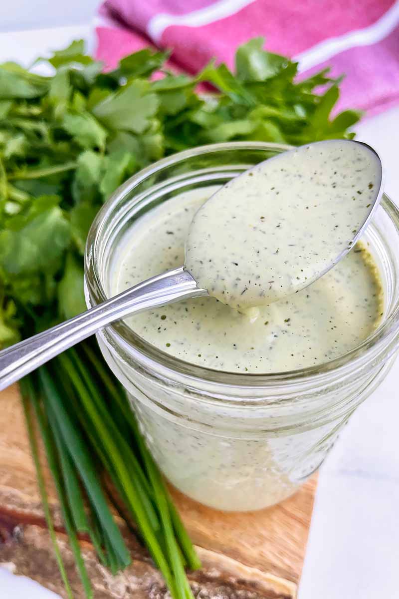 Vertical image of a spoon over a jar with a creamy herb condiment next to parsley.
