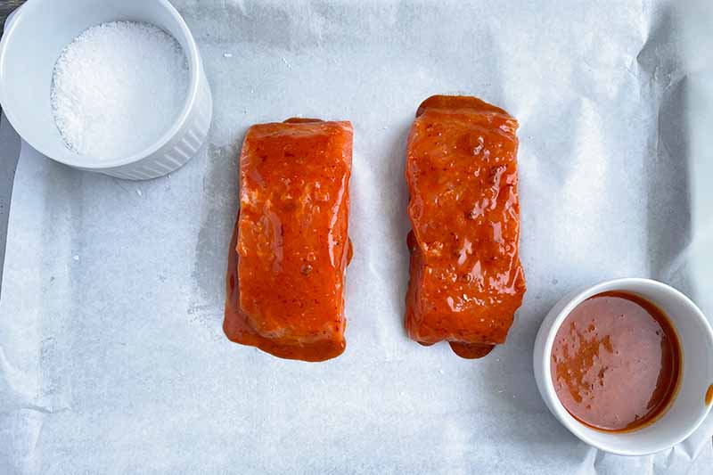 Horizontal image of two pieces of raw fish covered in a red sauce on a baking sheet lined with parchment paper next to ramekins of seasonings.