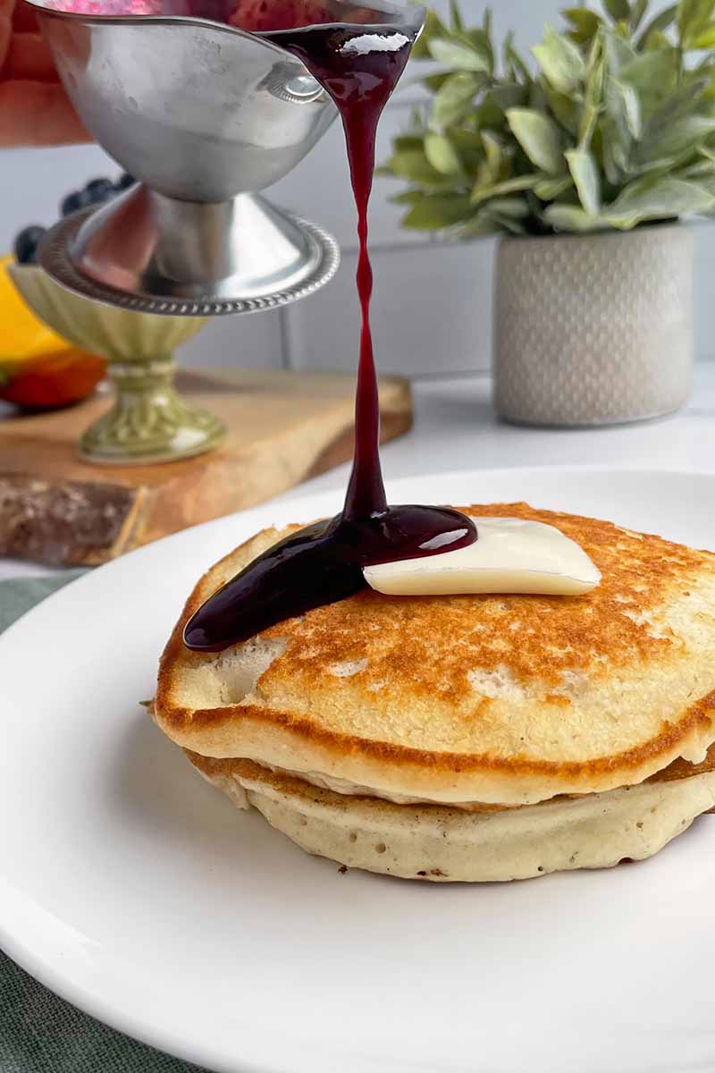 Vertical image of a dark purple syrup being poured on a stack of pancakes on a white plate.