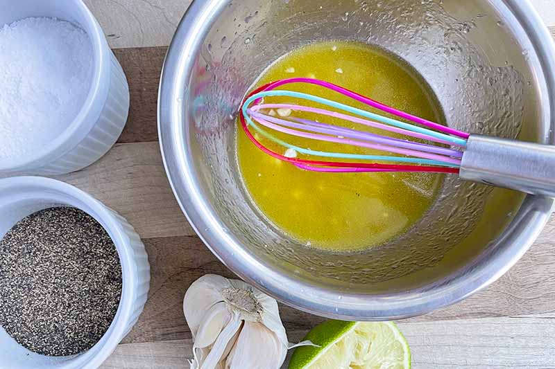Horizontal image of whisking a yellow vinaigrette in a metal bowl with seasonings on the side.