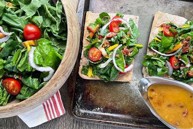 Horizontal image of pizza topped with greens and veggies on a sheet pan next to a big wooden bowl and dressing.