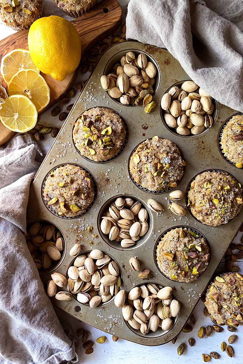 Vertical image of a tray full of muffins and pistachios next to slices of lemon.