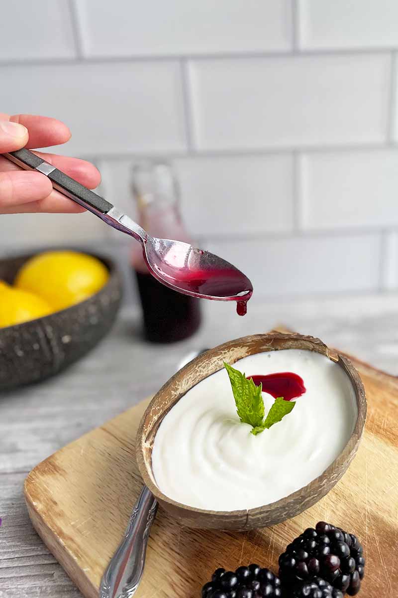 Vertical image of pouring a purple sauce on top of a wooden bowl filled with yogurt on a wooden cutting board.