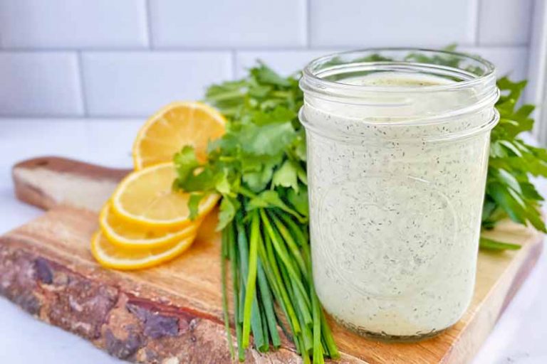 Horizontal image of a glass jar filled with an herb condiment next to lemons and parsley.