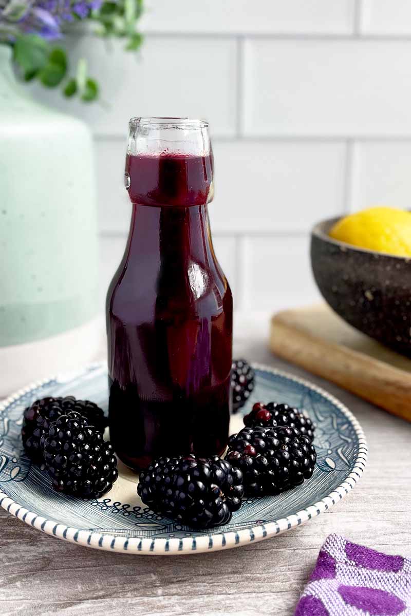 Vertical image of a tall glass bottle filled with a dark purple sauce on a small plate surrounded by whole blackberries.