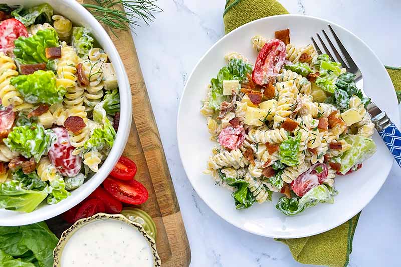 Horizontal image of a bowl and a plate with rotini, lettuce, and tomato in a creamy sauce.