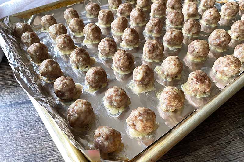 Horizontal image of rows of baked mini meatballs on a lined baking sheet.