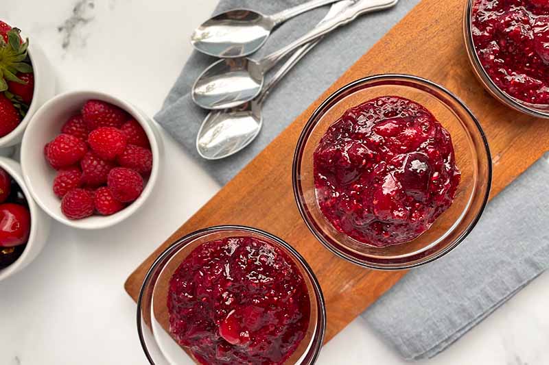 Horizontal image of three glass bowls filled with thick compote on a wooden plank next to silverware.