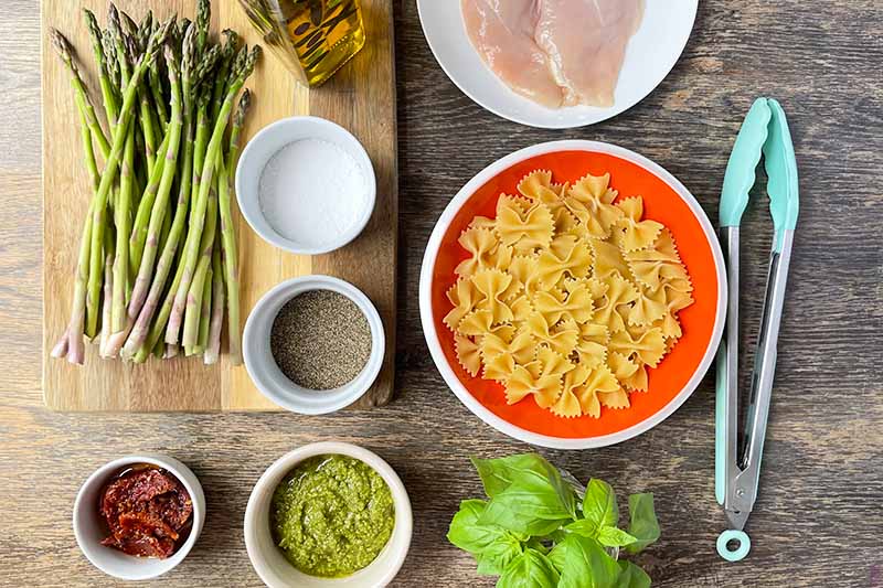 Horizontal image of various prepped ingredients, asparagus, farfelle, and raw poultry on a wooden table.