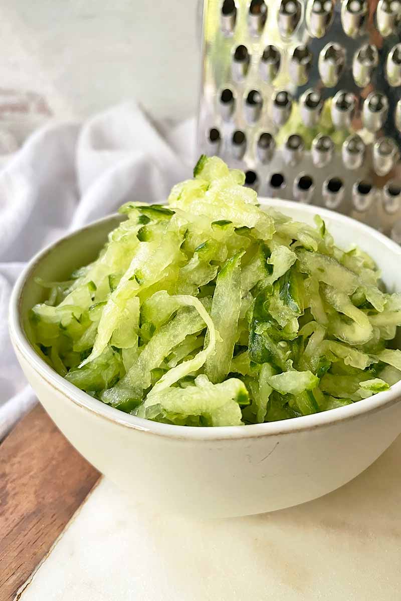 Vertical image of shredded cucumbers in a white bowl next to a metal kitchen tool.