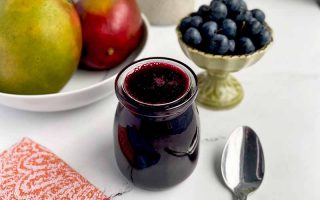 Horizontal image of a glass jar filled with a dark purple sauce next to a spoon, fresh fruit, and a colorful napkin.