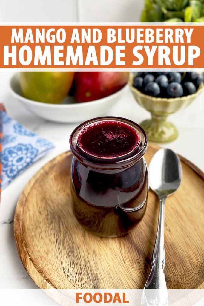 Vertical image of a glass jar filled with a purple sauce on a wooden plate next to silverware and fresh fruit.