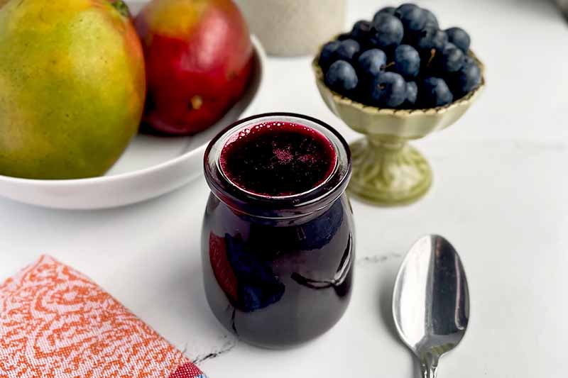 Horizontal image of a glass jar filled with a dark purple sauce next to a spoon, fresh fruit, and a colorful napkin.