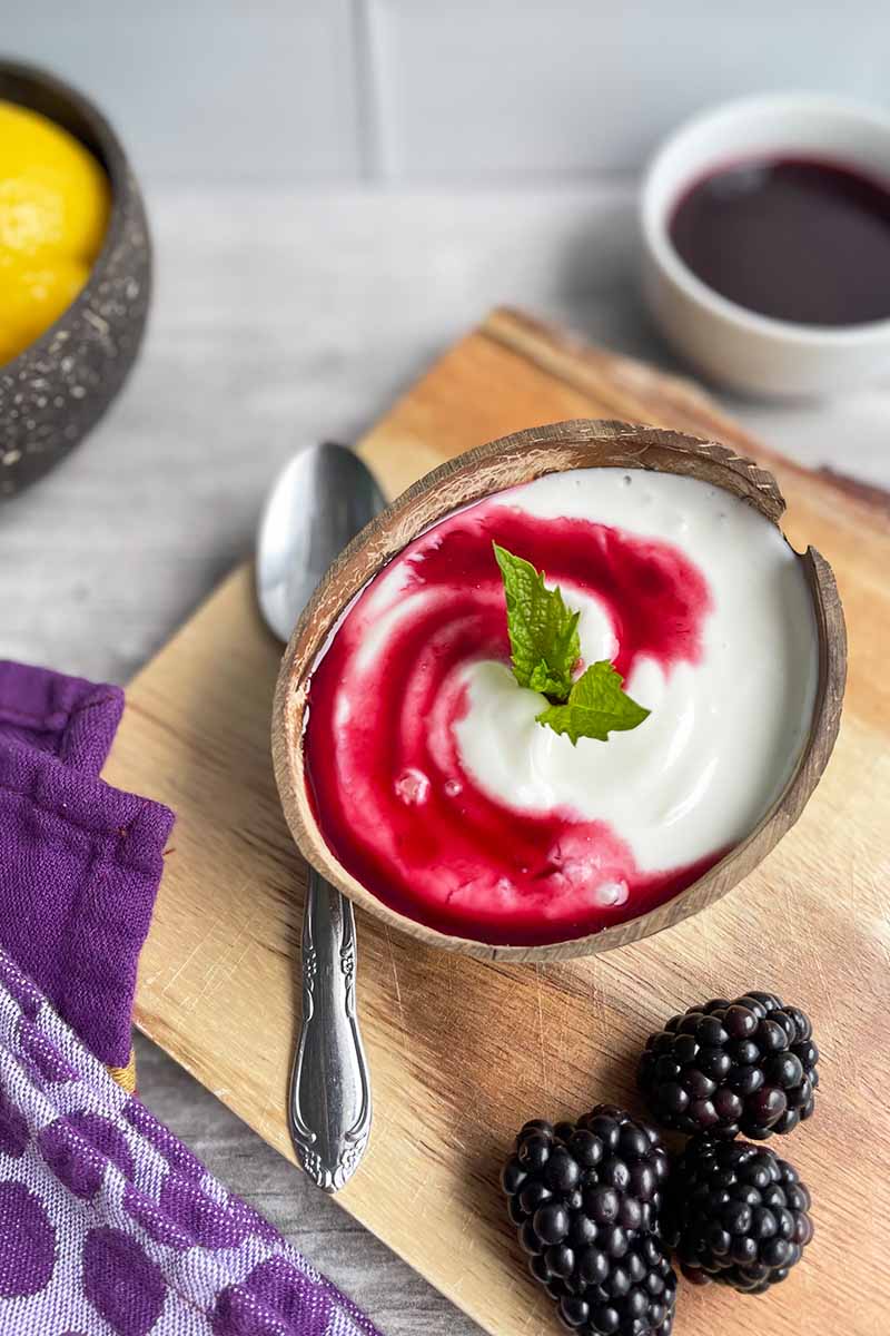 Vertical image of a wooden bowl filled with plain yogurt topped with a blackberry sauce and mint leaves, on a wooden board next to a spoon and fresh fruit.