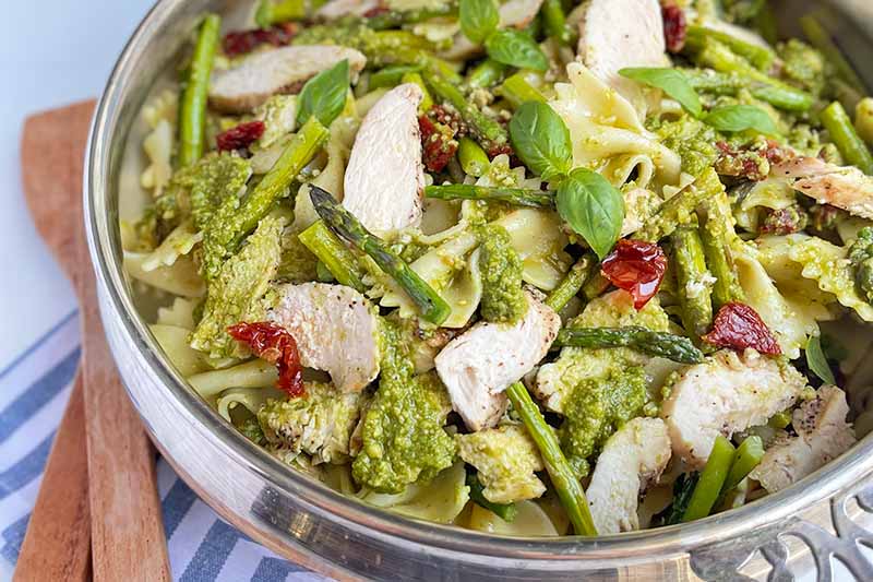 Horizontal image of a large bowl filled with a pasta dish with green sauce, vegetables, and sliced cooked poultry.